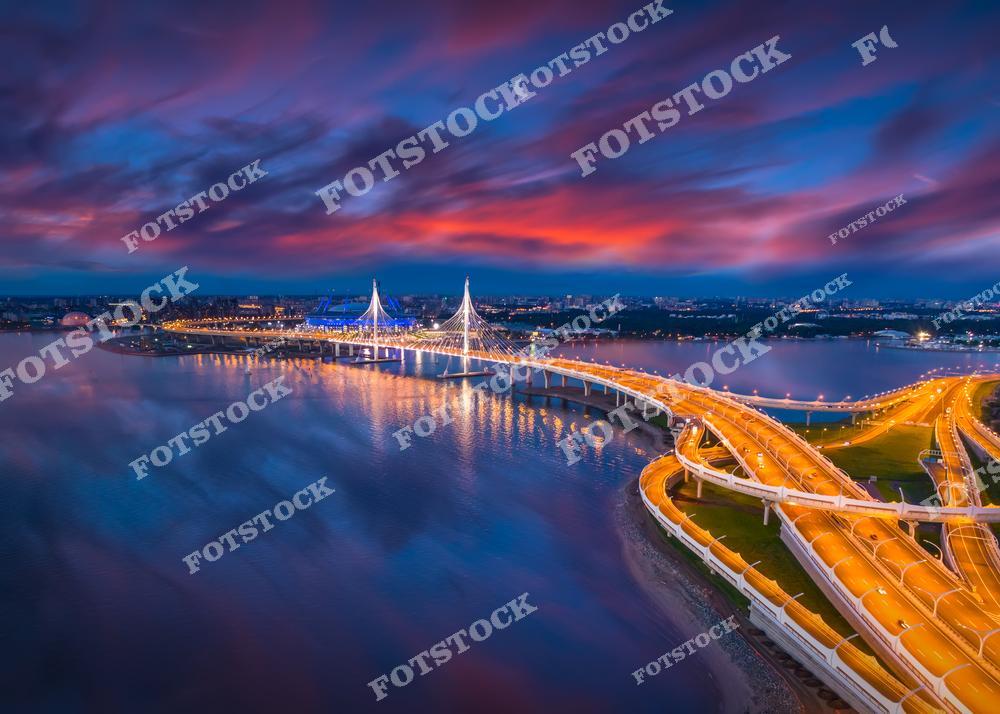 На фотографии запечатлен Западный скоростной диаметр (ЗСД) в вечернее время, проходящий над Финским заливом. Мост и трасса освещены, их линии плавно уходят в перспективу, создавая живописный вид на фоне ярких красок заката.